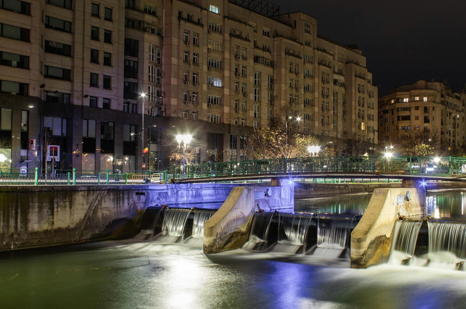 A lit stepped weir on a city river at night flanked by apartment blocks