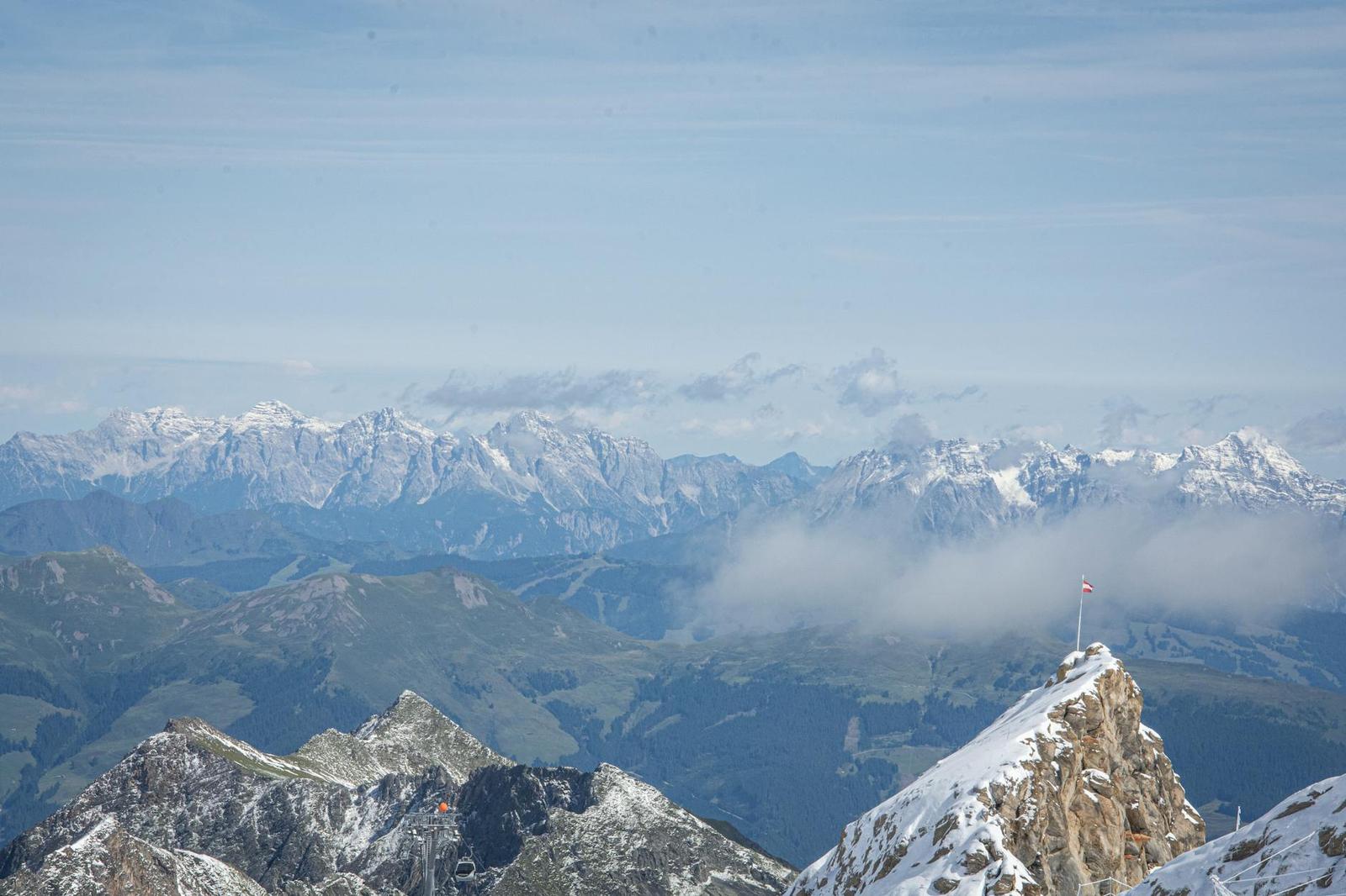 A small flag marking a rocky summit above layered snow-capped mountain ranges