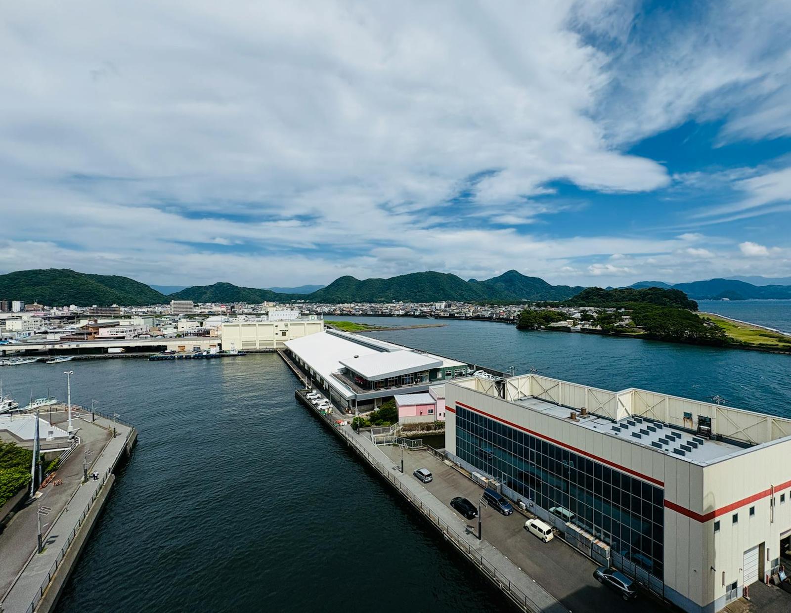 Aerial view of a waterfront building complex beside a harbour with hills behind