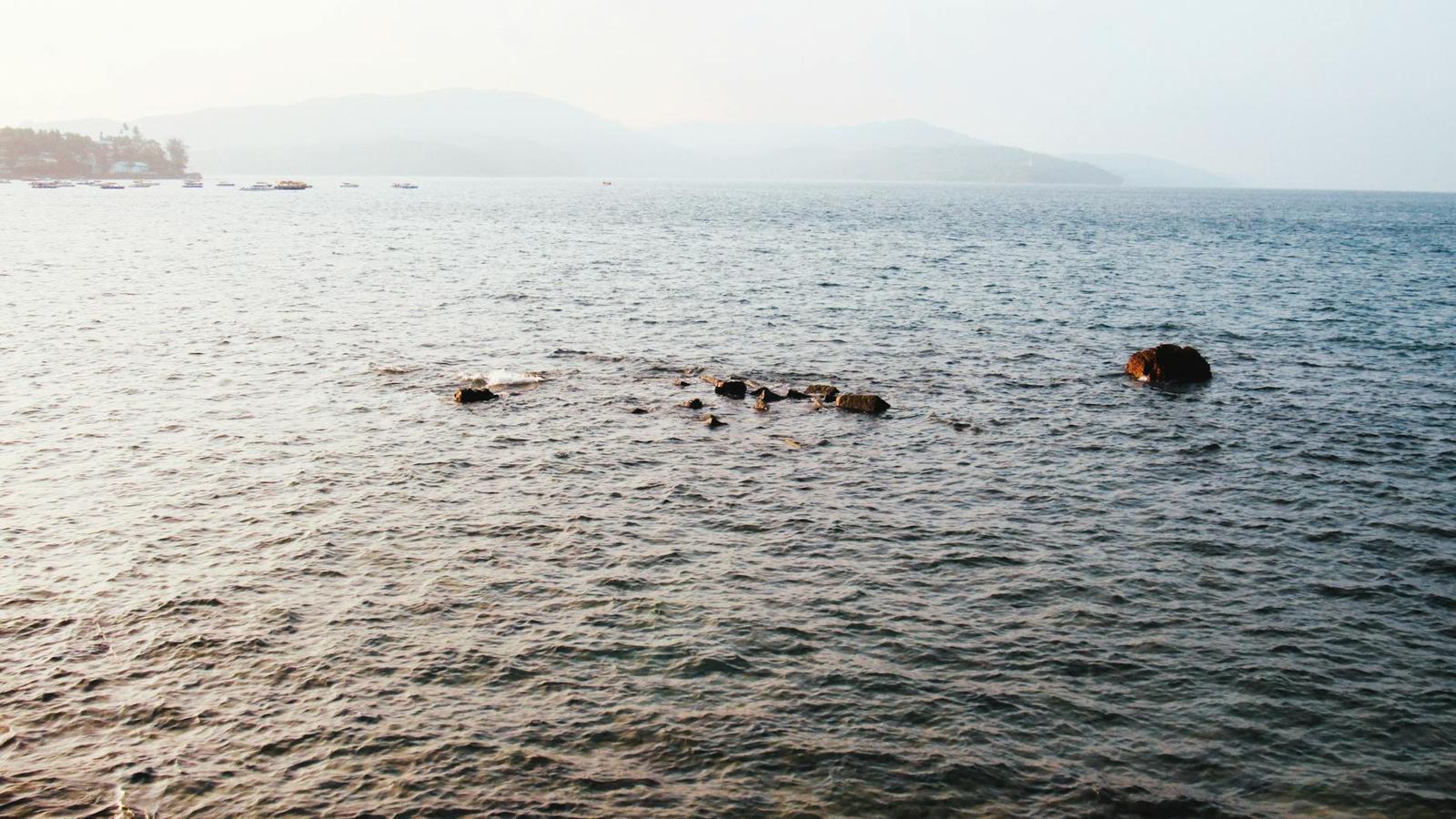 Dark rocks breaking the surface of a hazy sea with distant hills
