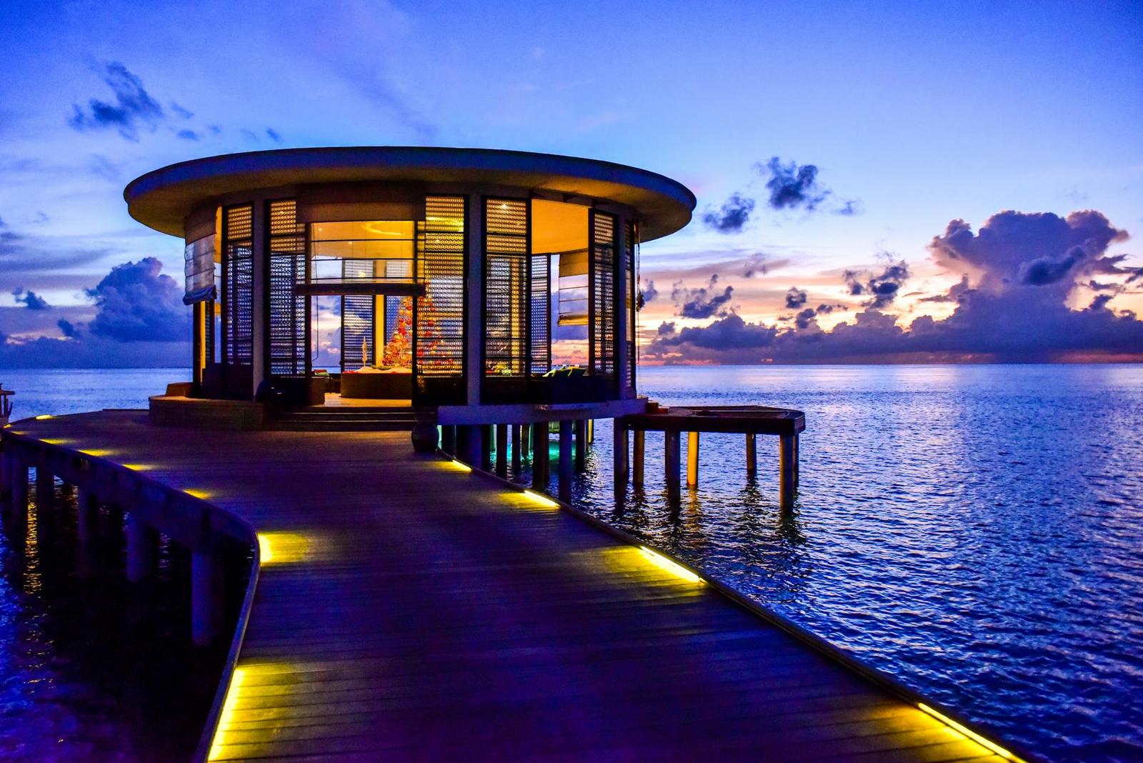 A circular glass-walled overwater lounge lit up at dusk