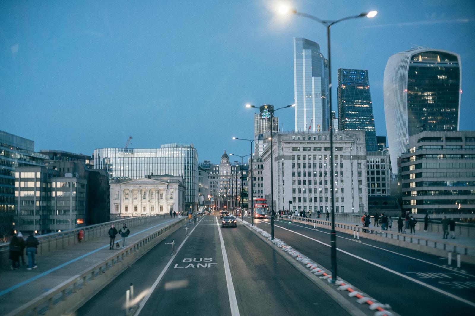 A city bridge at dusk with pedestrians walking beside traffic and lit towers ahead