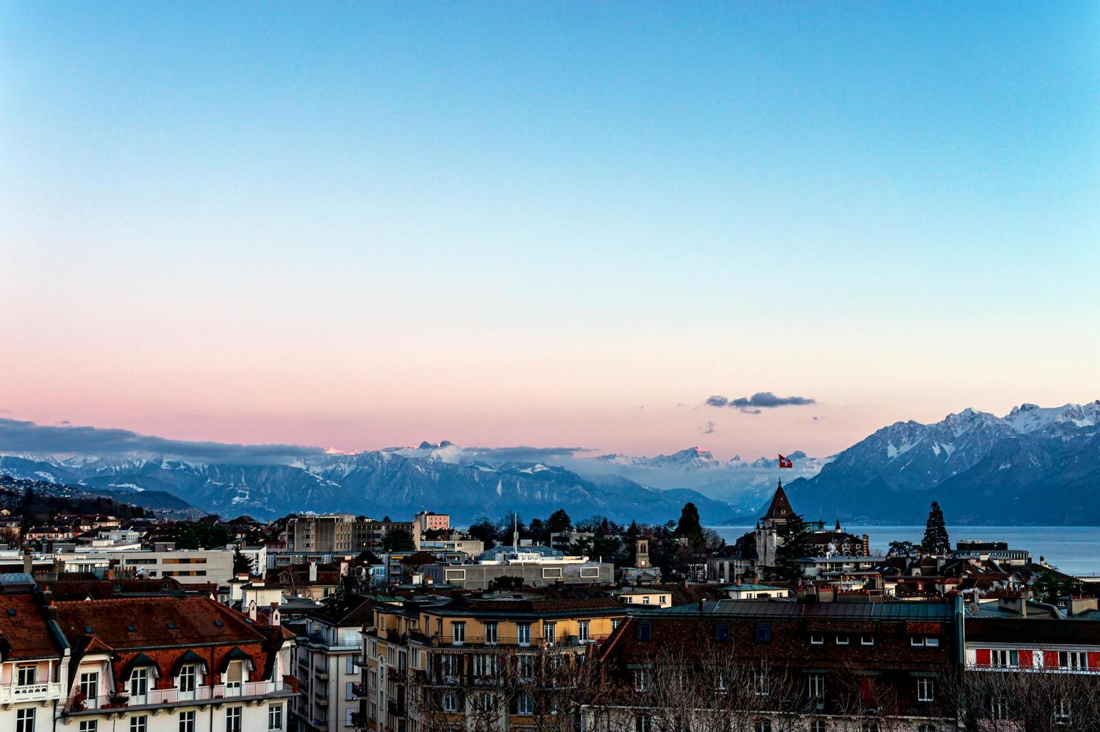 City rooftops silhouetted against a pastel dusk sky with lake and mountains