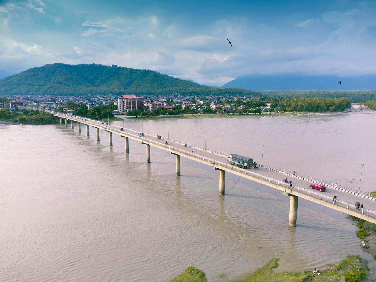 A long road bridge spanning a wide muddy river beneath green hills