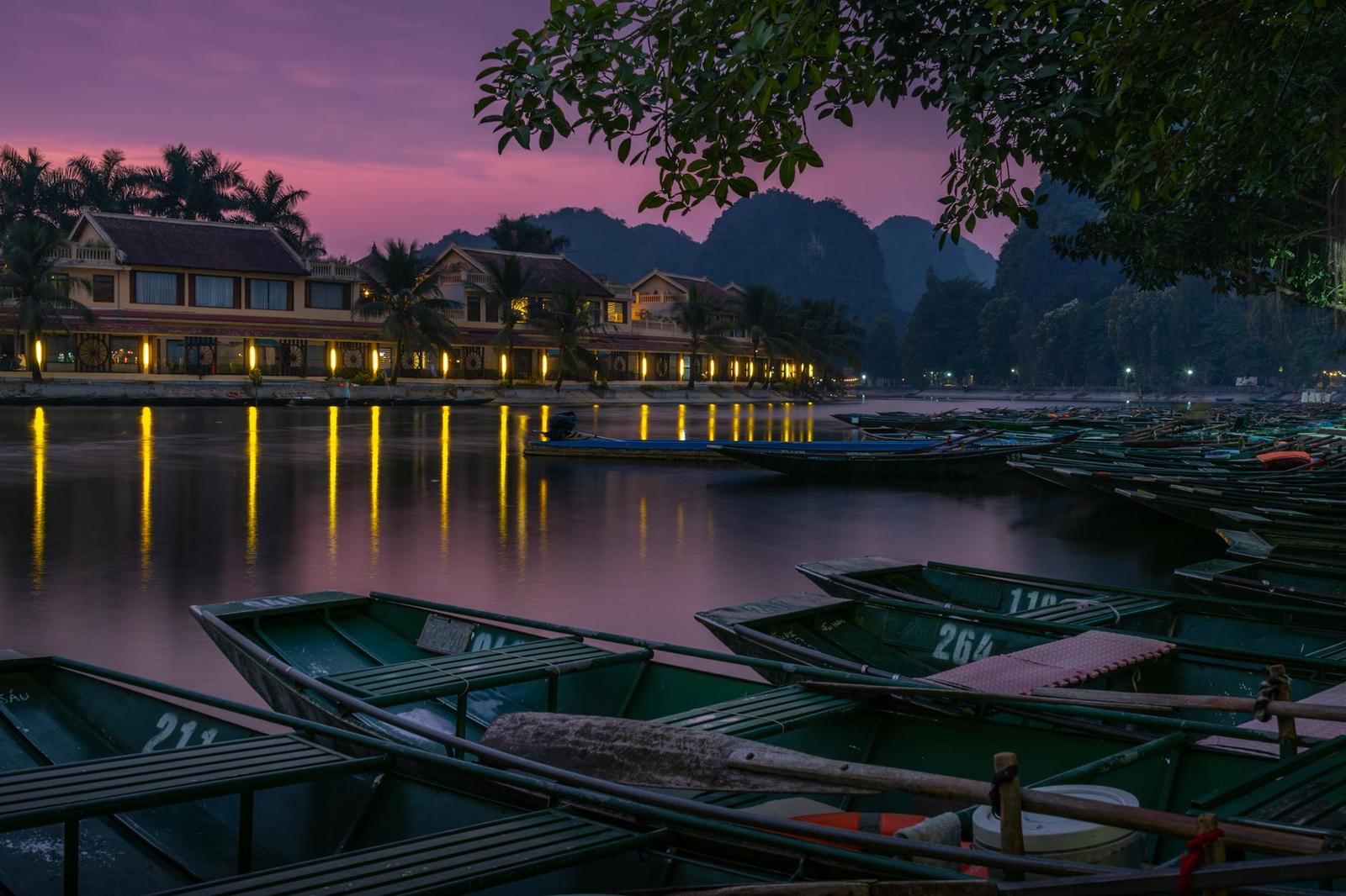 Rowboats moored at dusk beside a riverside village lit against a purple sky