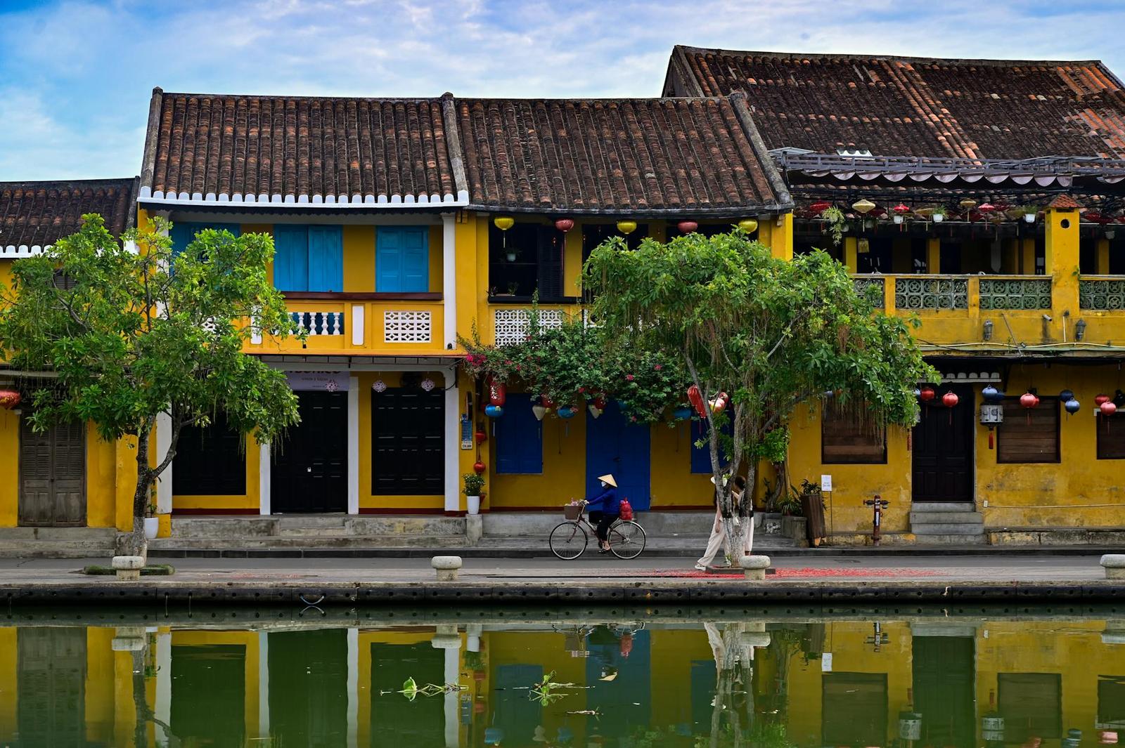 A yellow shophouse row with red lanterns and a cyclist reflected in a canal