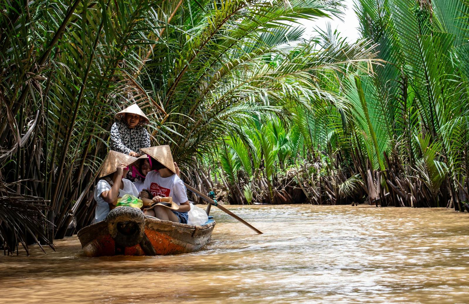 Passengers in conical hats riding a wooden boat through a palm canal