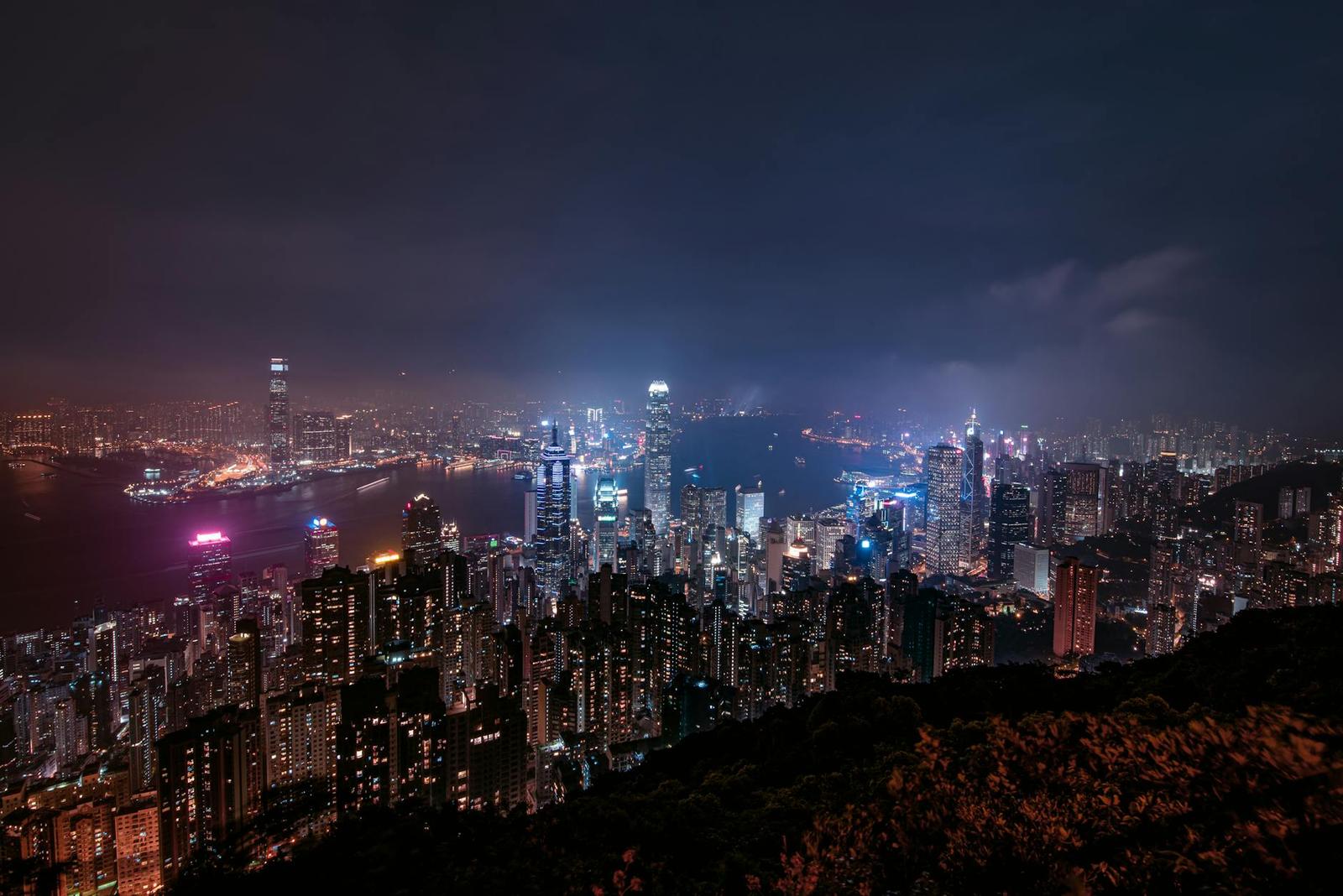 A dense high-rise skyline lit up at night around a harbour, seen from a hilltop