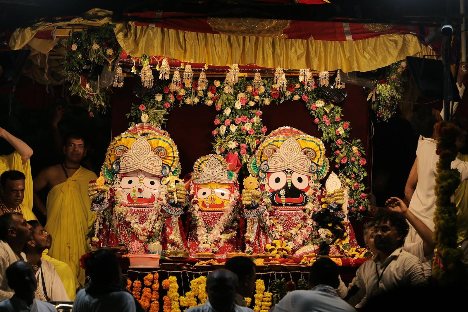 Three garlanded temple idols decorated with flowers beneath a canopy at night