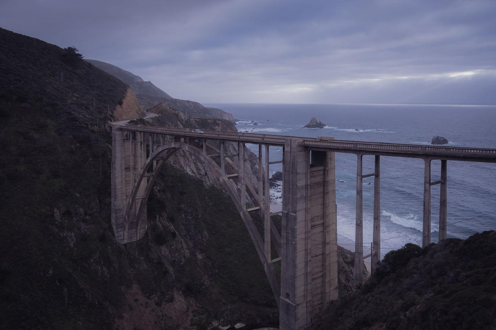 An arched concrete bridge over a rocky coastline under a cloudy dusk sky
