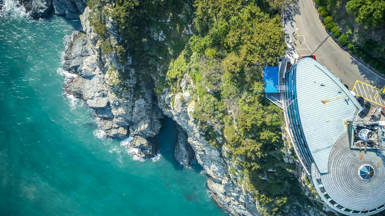 Aerial view of a rocky cove with turquoise water beside a curved blue-roofed building