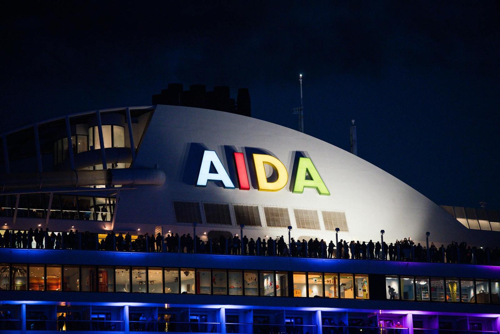 A brightly lettered cruise ship hull glowing at night with passengers lining the deck