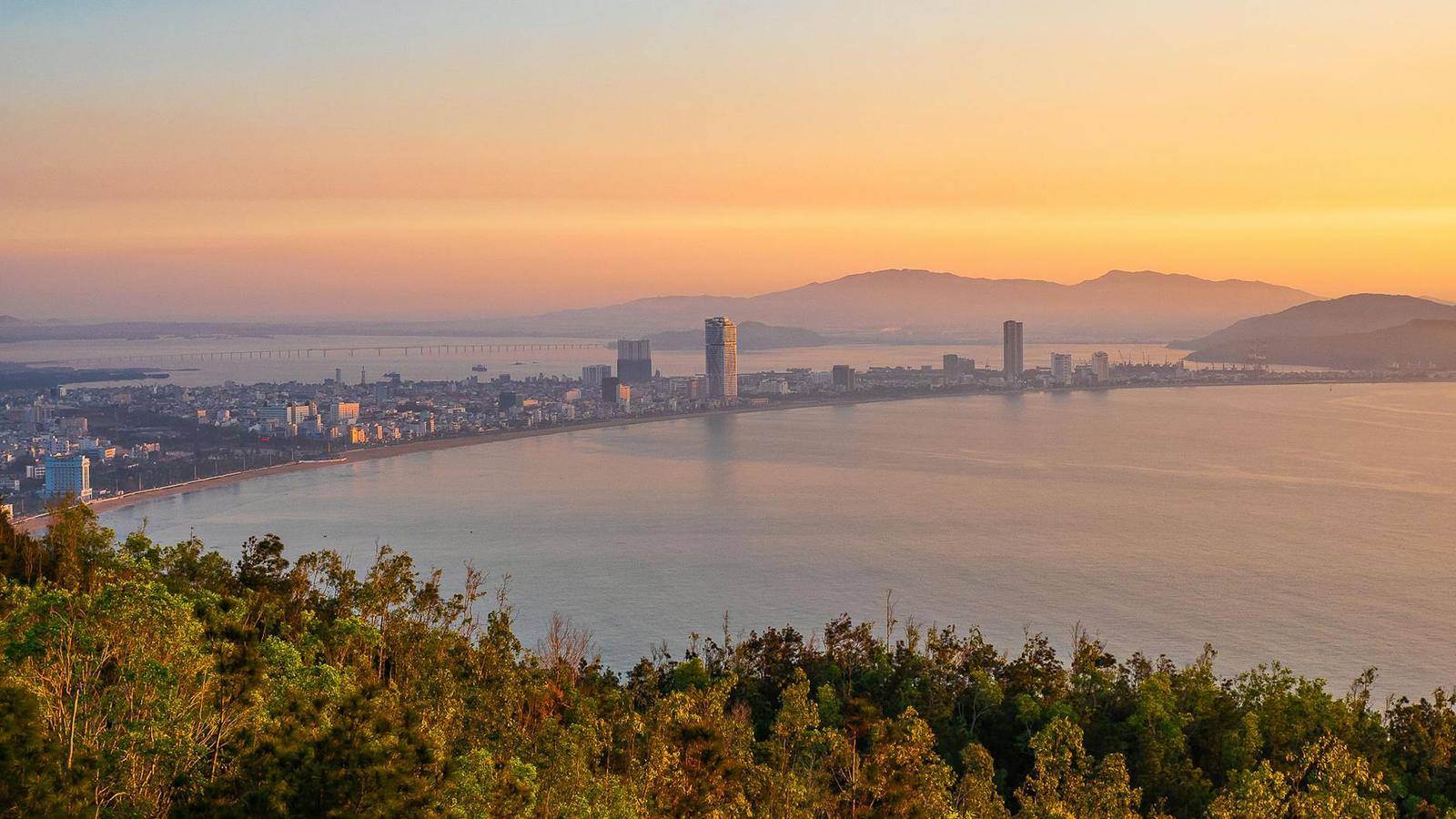Coastal city skyline silhouetted against a bay at sunset seen from a forested hill