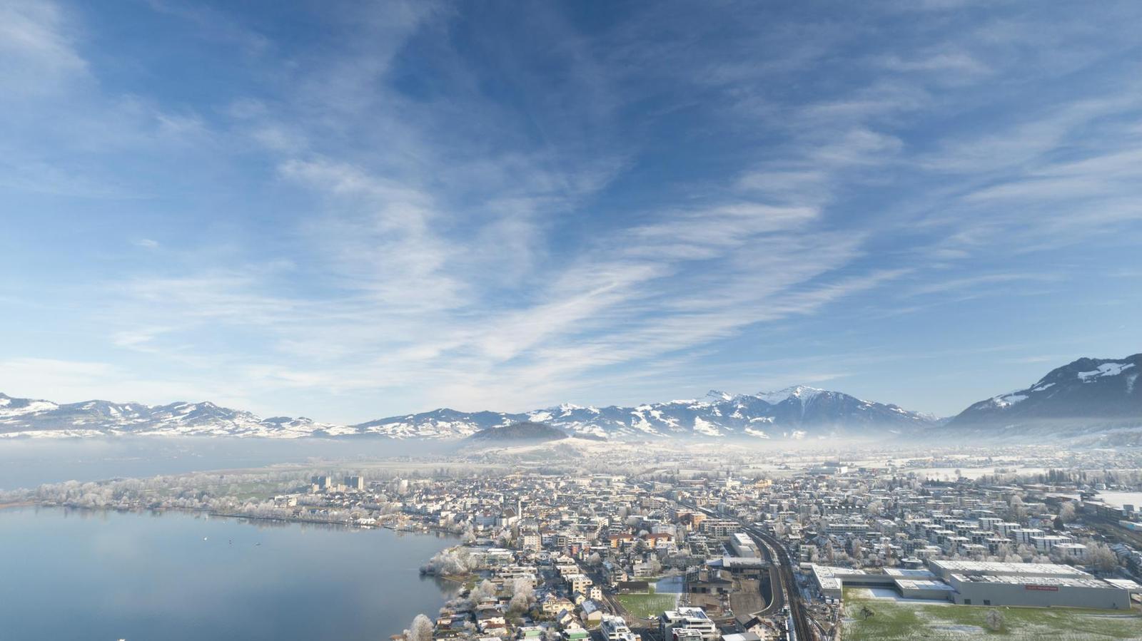 Aerial view of a lakeside city with mist rising toward snow-dusted mountains