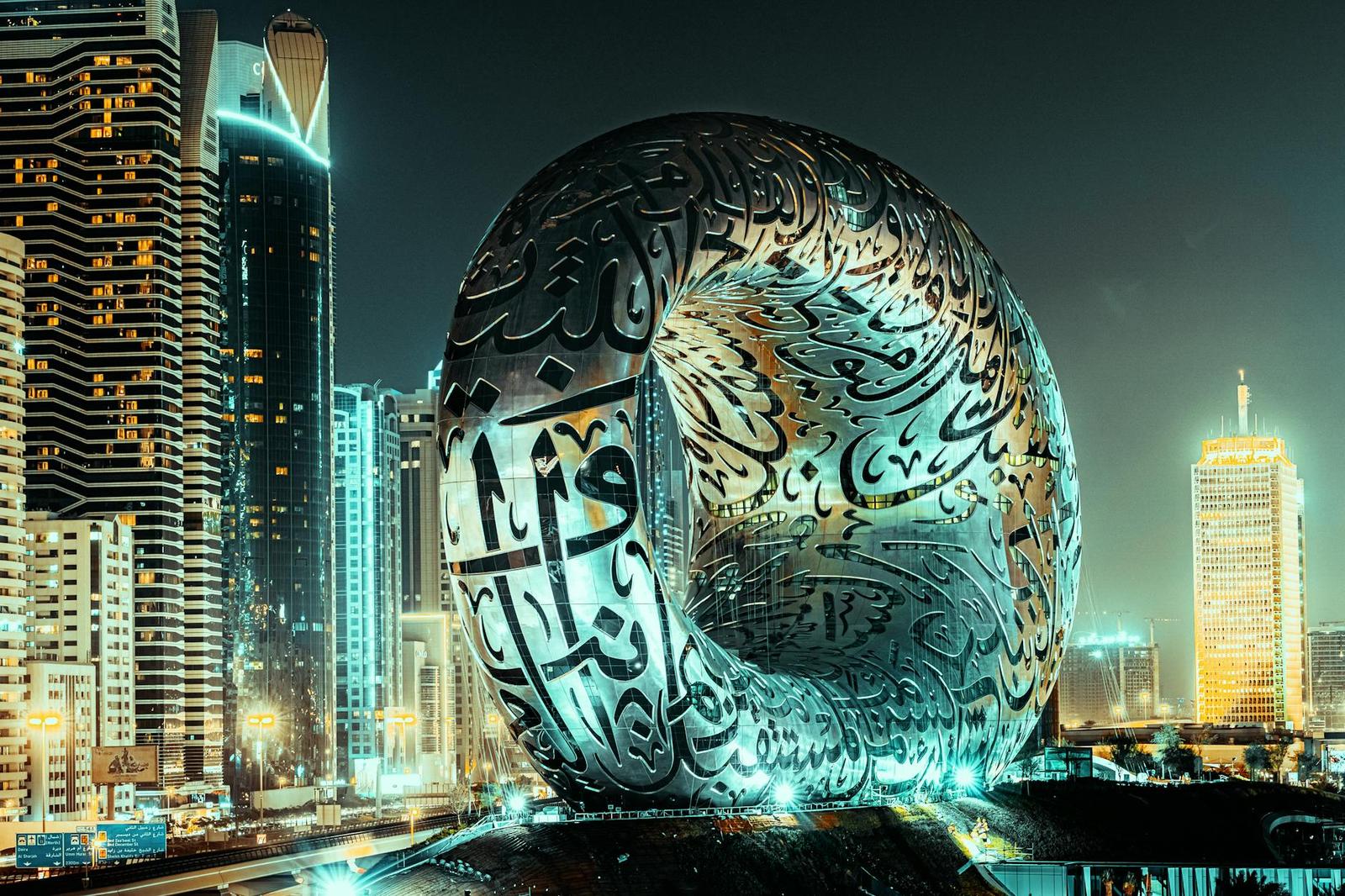 An oval building covered in calligraphy script lit up at night among city towers