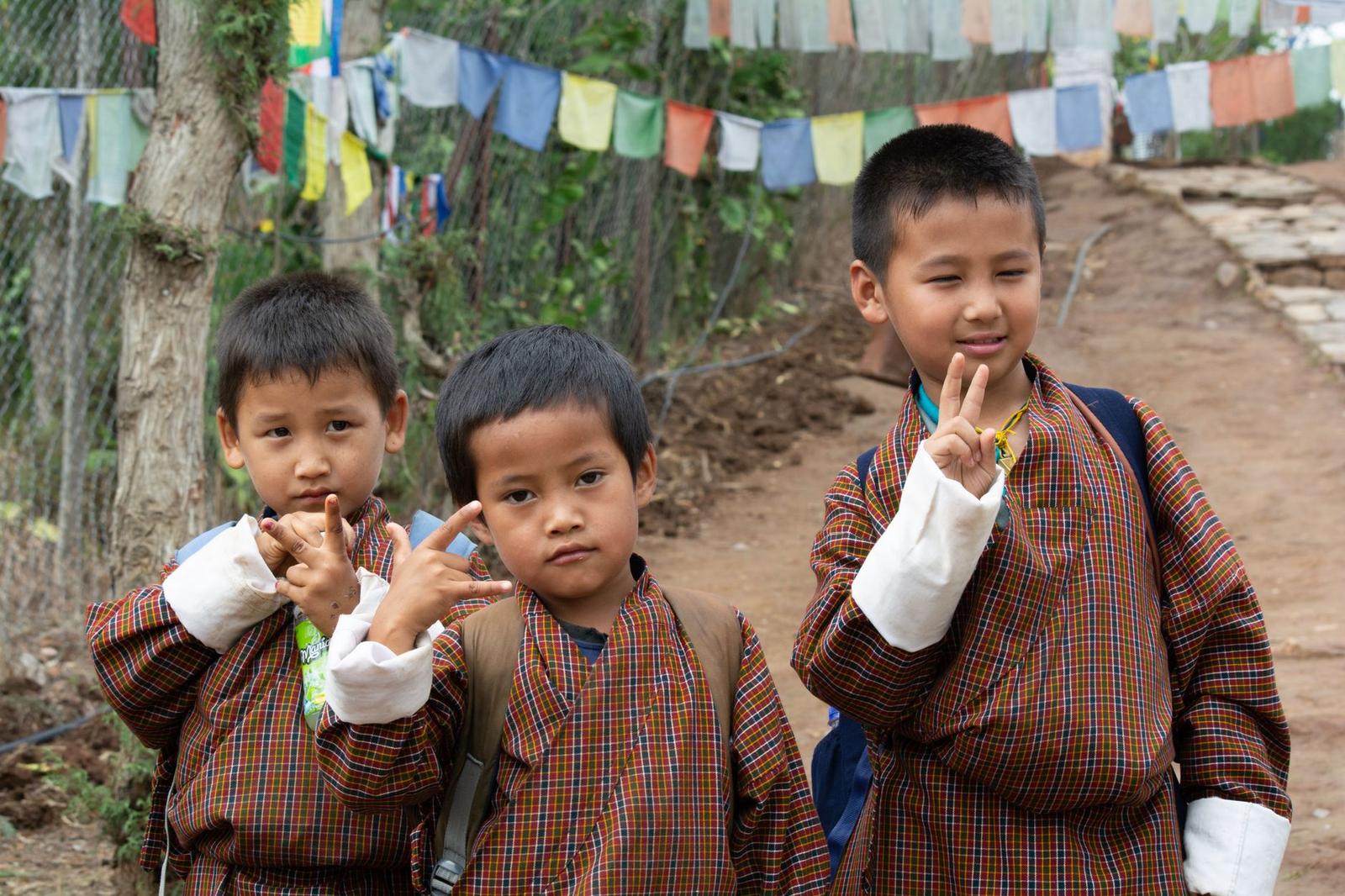 Three boys in traditional dress making peace signs beneath prayer flags