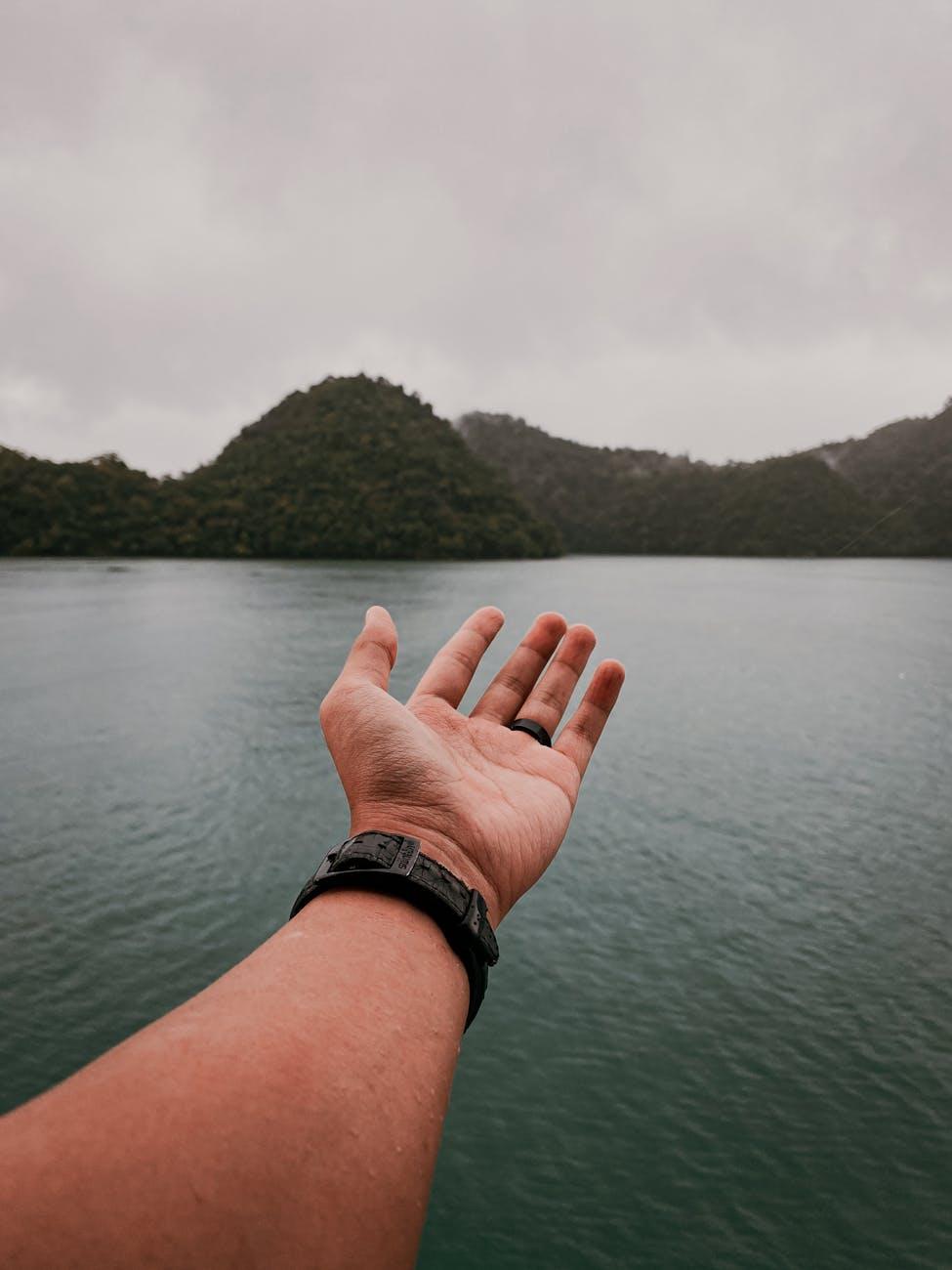 Outstretched hand reaching toward a still lake framed by forested hills