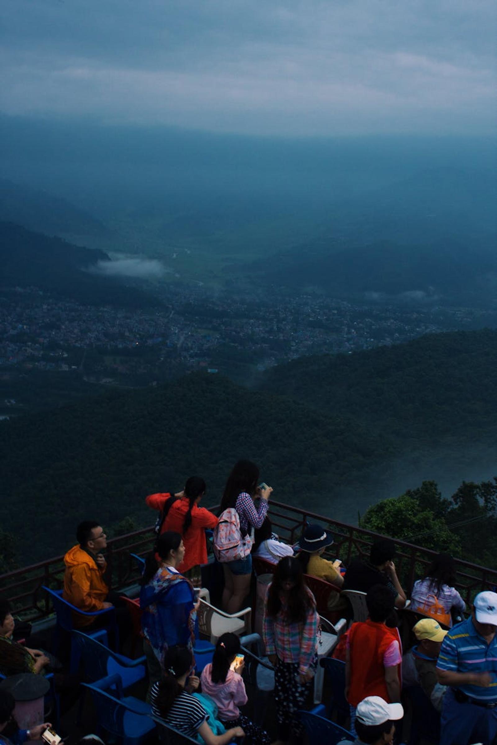 A crowd gathers at a scenic lookout over Pokhara, Nepal, with misty mountains in the distance