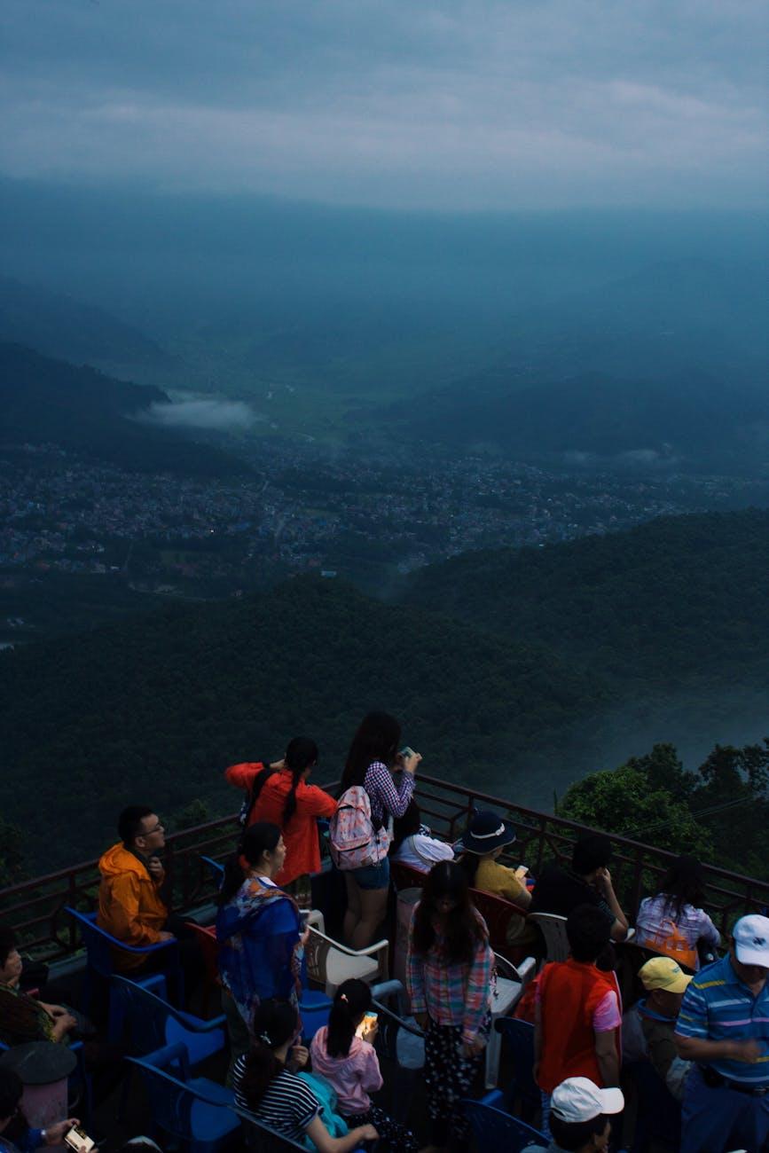 A crowd gathers at a scenic lookout over Pokhara, Nepal, with misty mountains in the distance