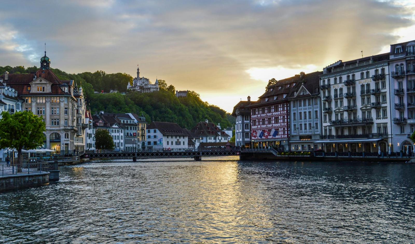 Riverside townhouses glowing under a golden evening sky with a hillside chapel behind
