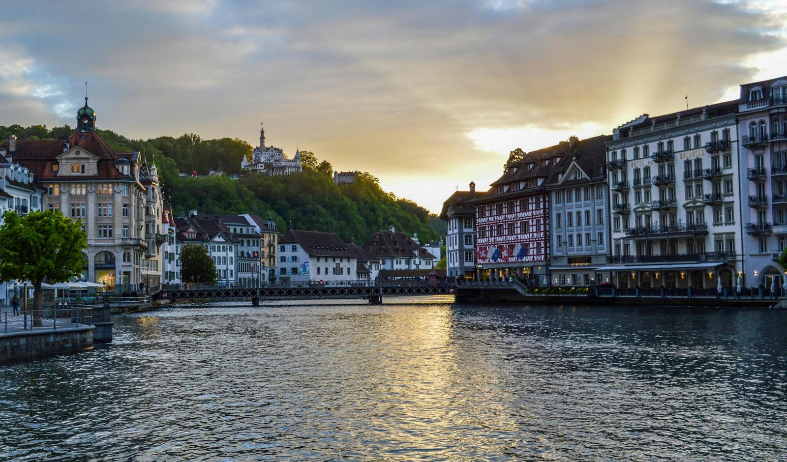 Riverside townhouses glowing under a golden evening sky with a hillside chapel behind