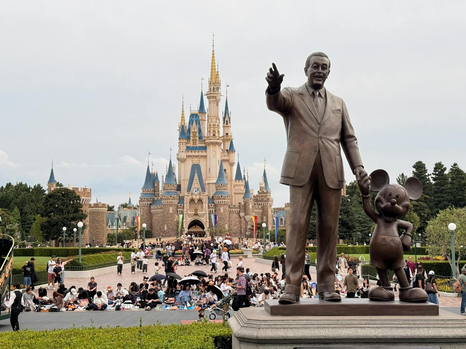 A bronze statue of a man and a mouse character before a fairytale castle with crowds gathered