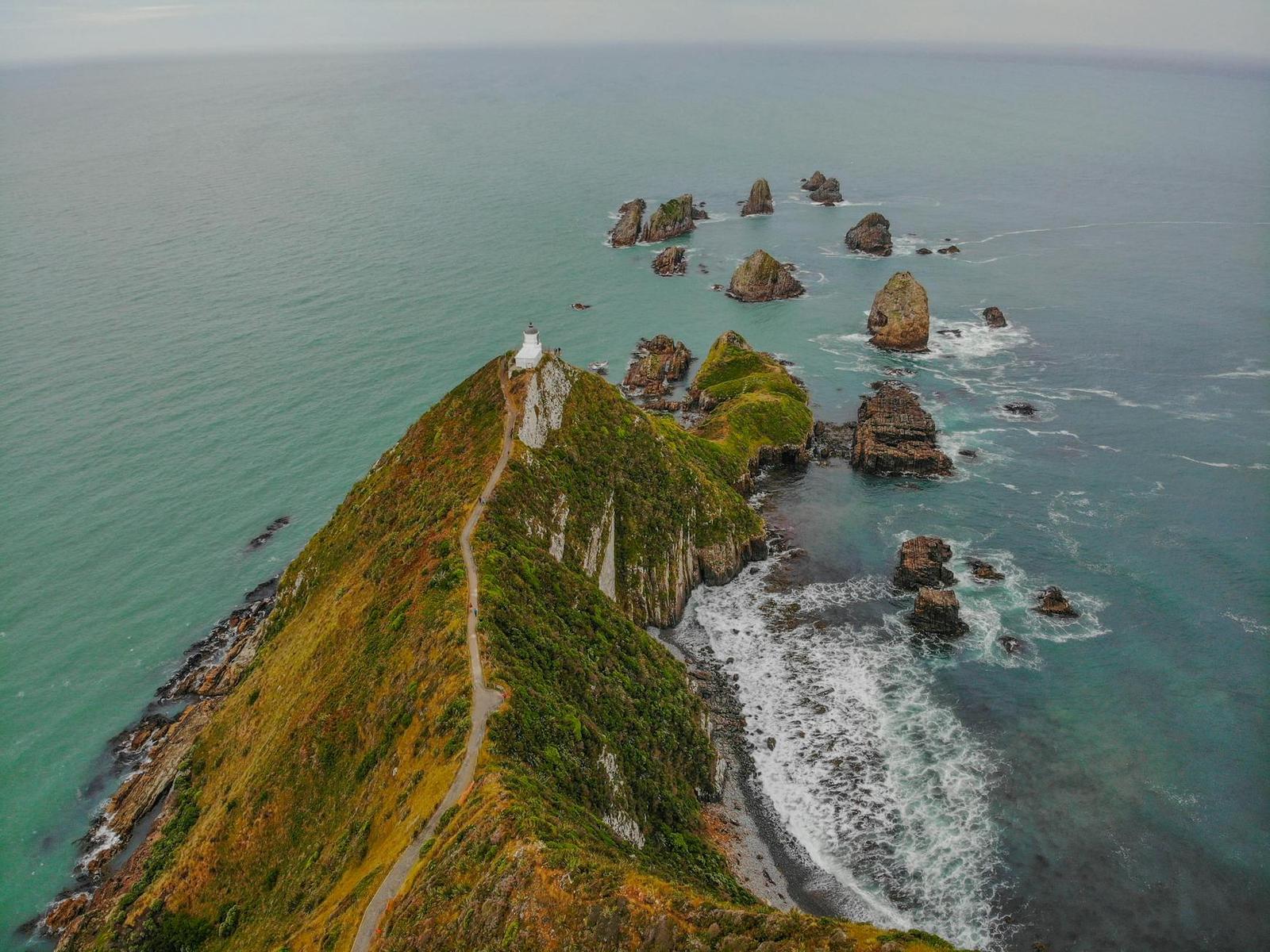 Aerial view of a grassy headland and lighthouse jutting into turquoise sea