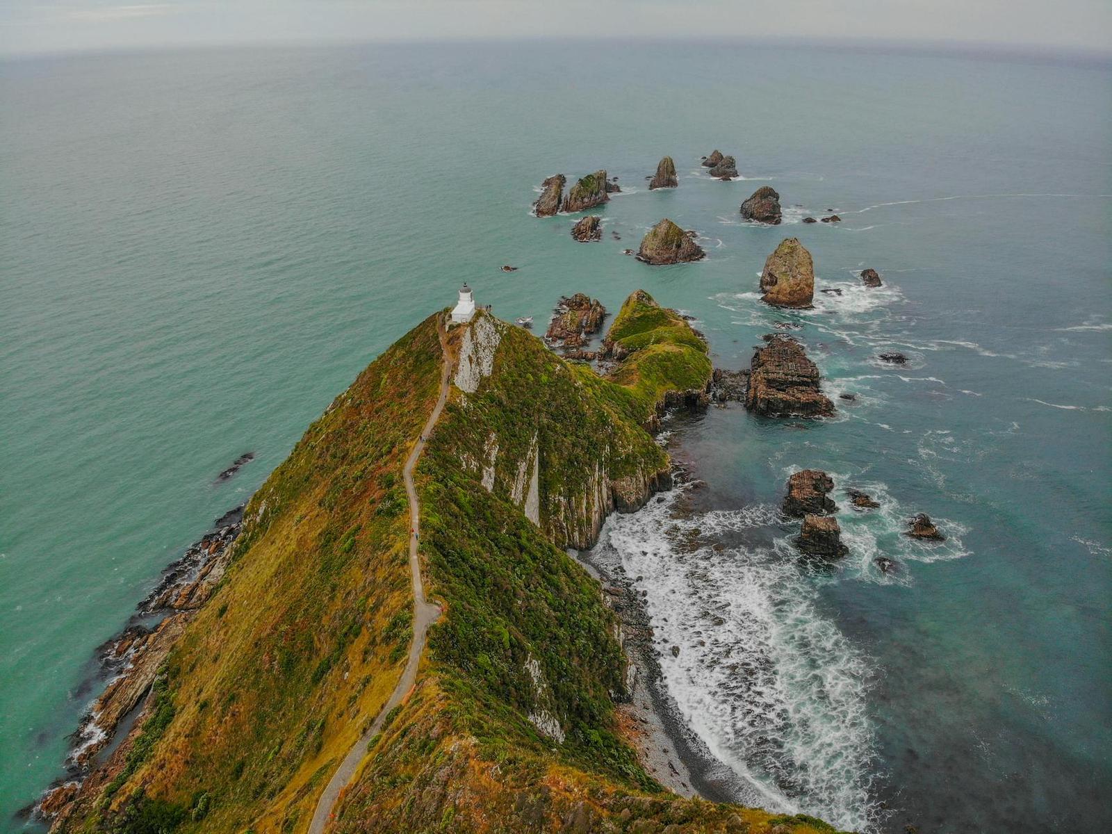 Aerial view of a grassy headland and lighthouse jutting into turquoise sea