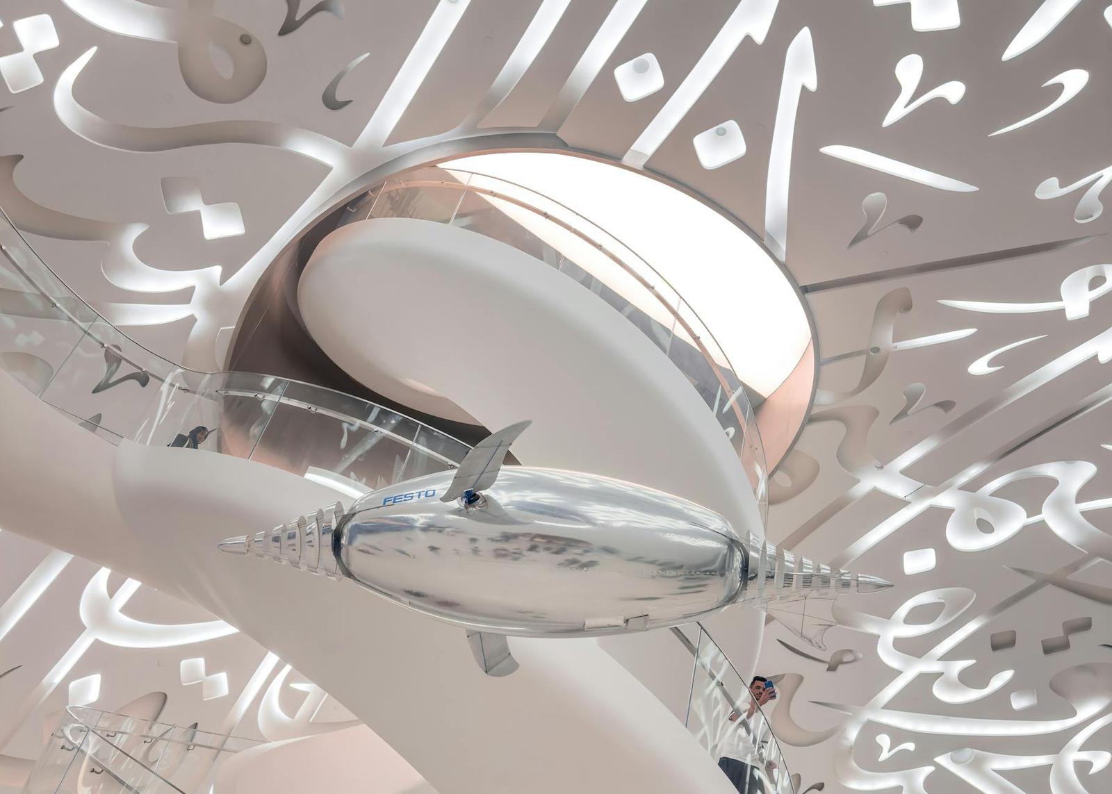 A curved white gallery interior with flowing script carved into the ceiling and a lone visitor below