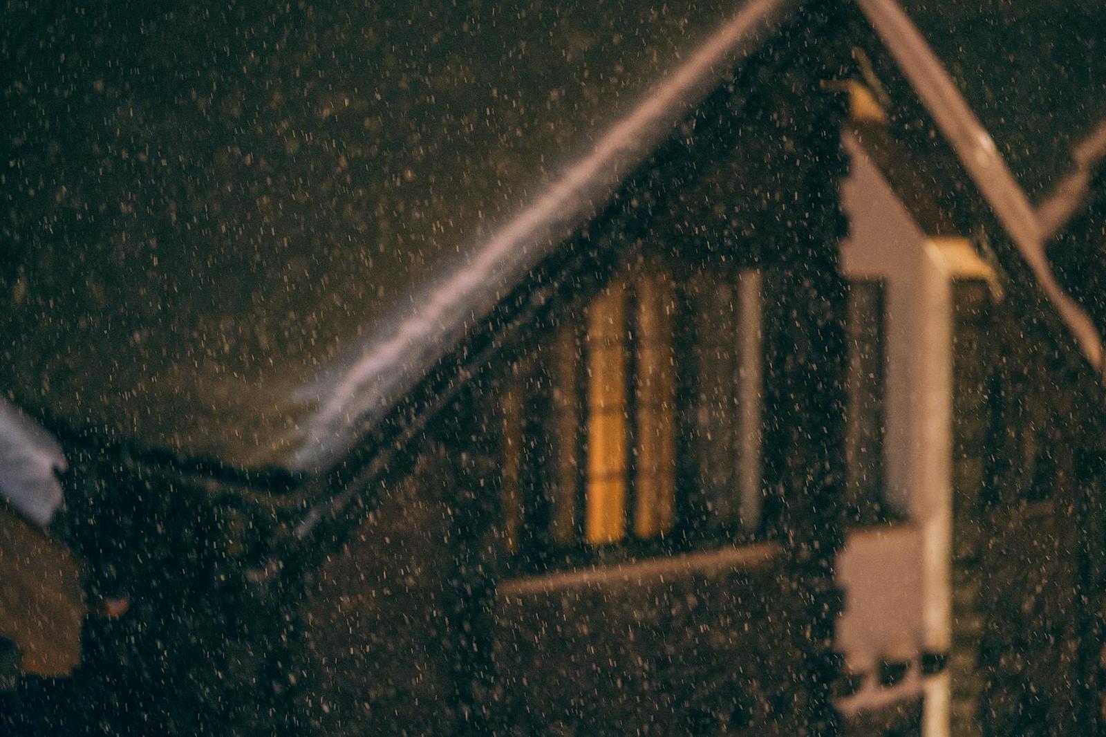 A wooden gabled house lit from within during heavy snowfall at night