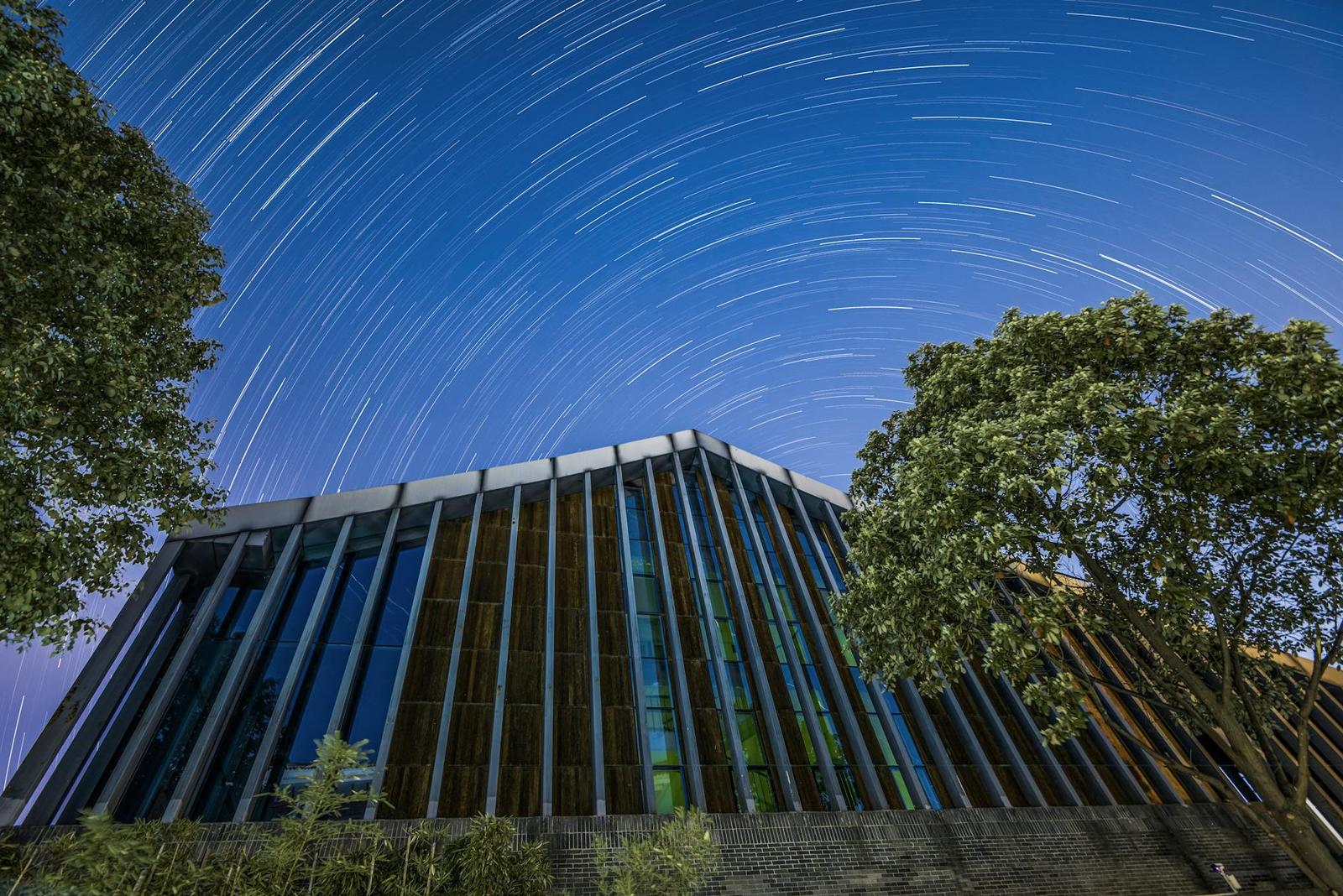 Star trails circle above an angular building framed by trees at night