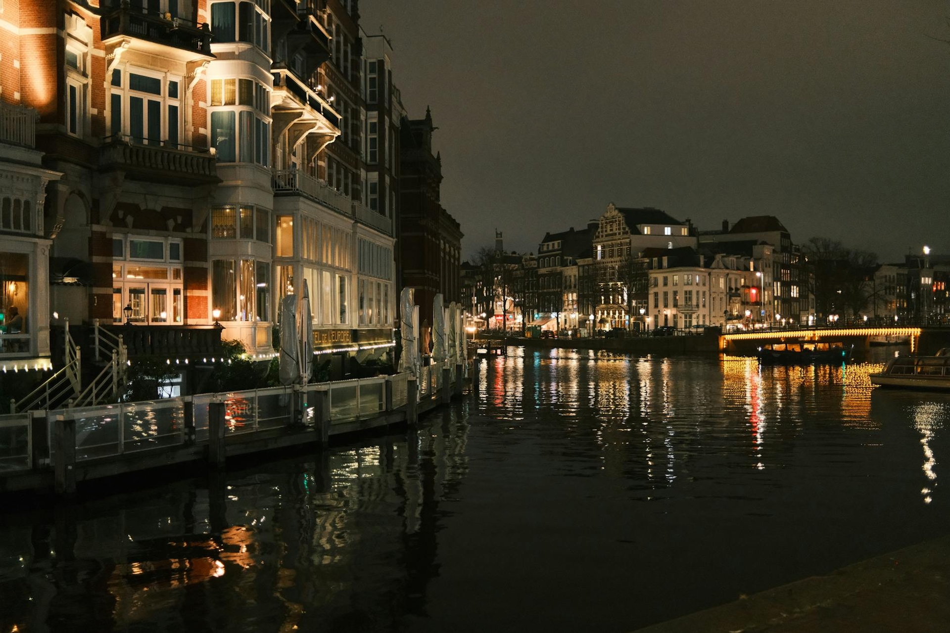 A lit riverside hotel terrace overlooking a dark canal with a bridge beyond