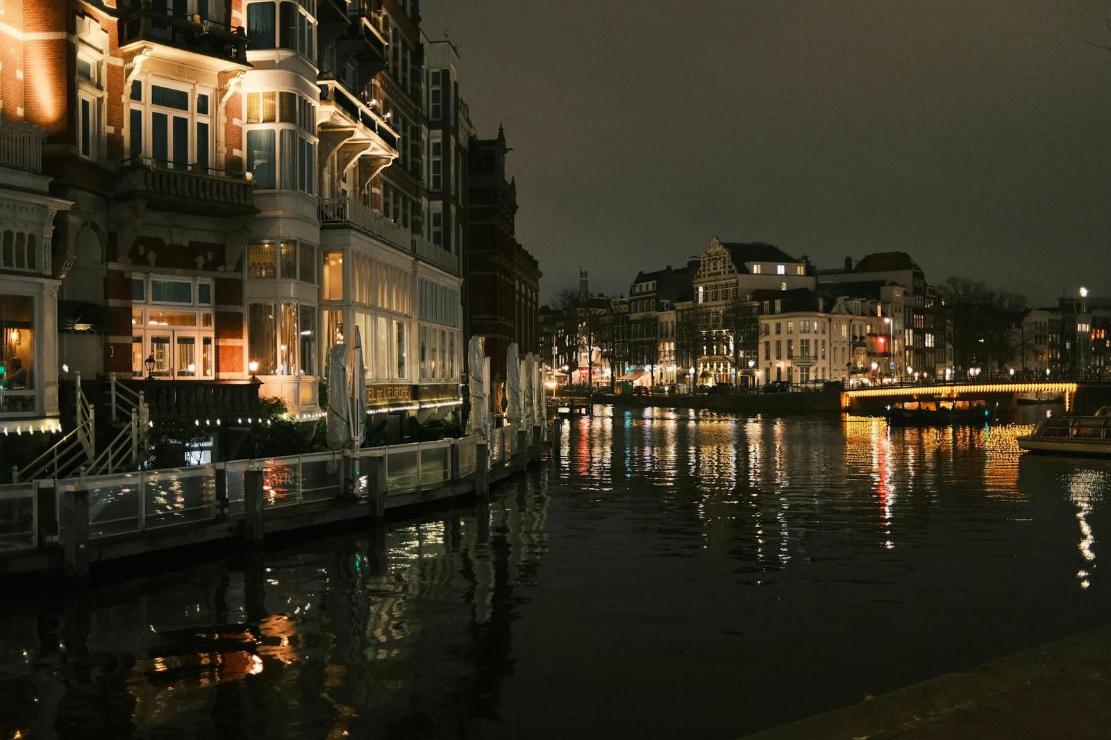 A lit riverside hotel terrace overlooking a dark canal with a bridge beyond