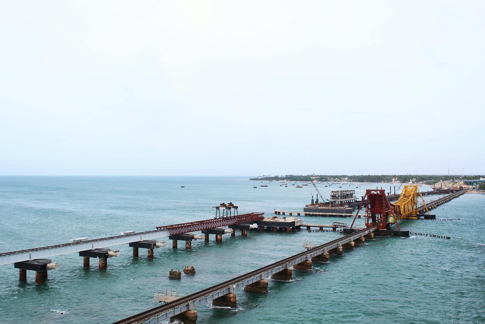A long railway bridge with cranes extending across calm coastal water