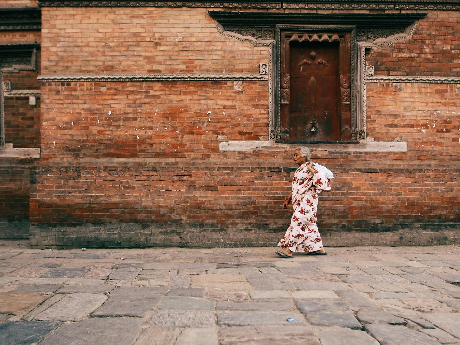 A woman in a floral sari walking along a cobbled street past a brick temple wall