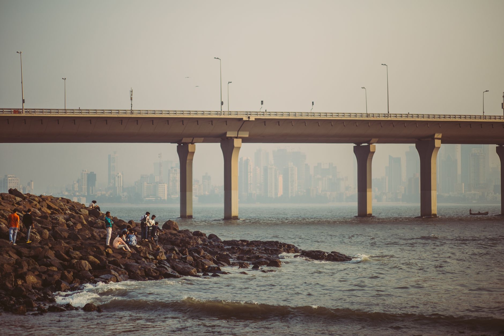 People fishing from a rocky seawall beneath a long coastal bridge