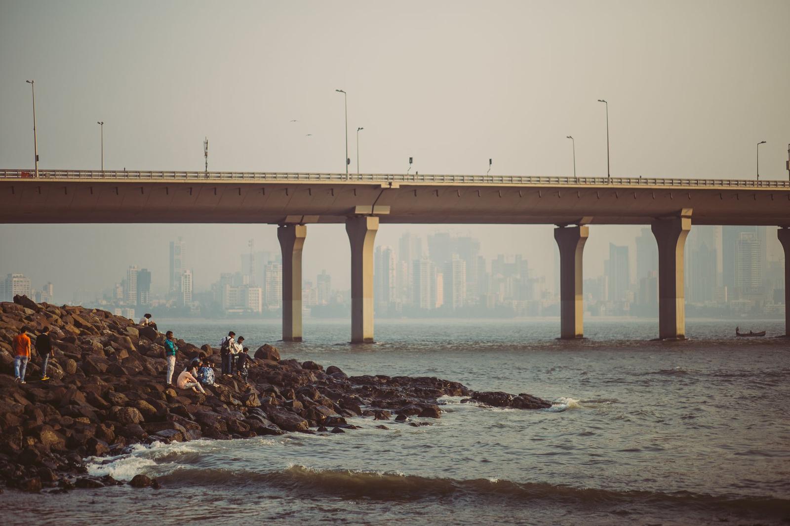 People fishing from a rocky seawall beneath a long coastal bridge