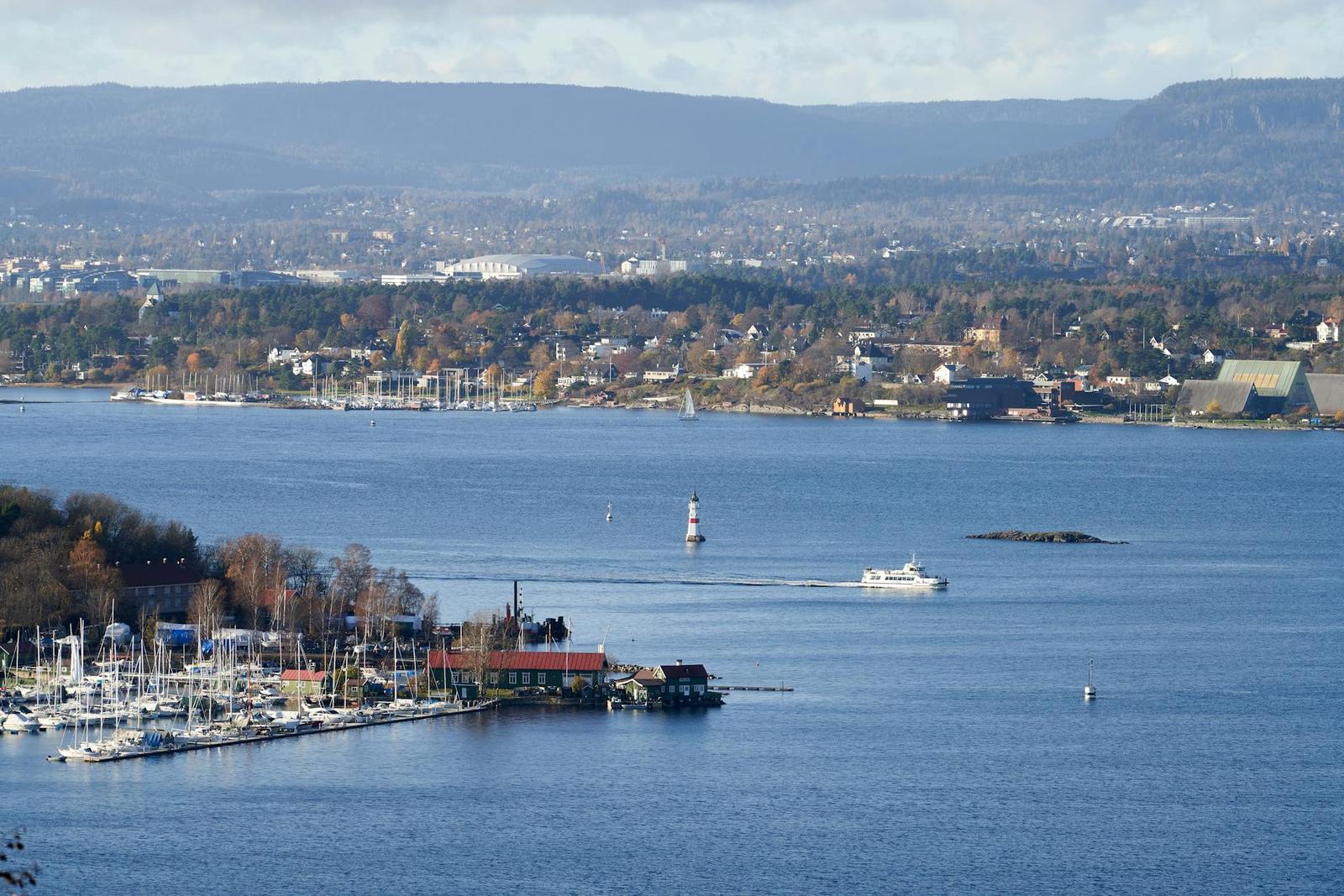 A small boat crossing a wide fjord past a marina and a red lighthouse buoy