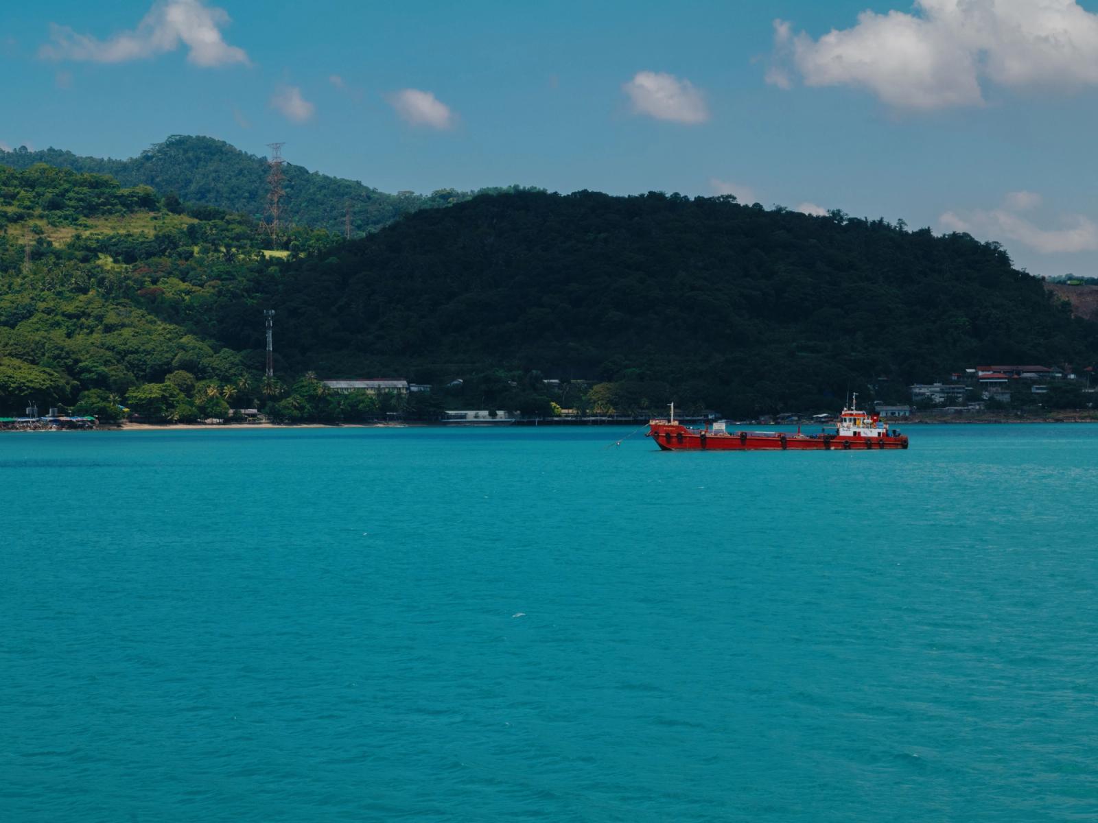 A red ship sails the vibrant blue waters of a tropical island coastline in Indonesia