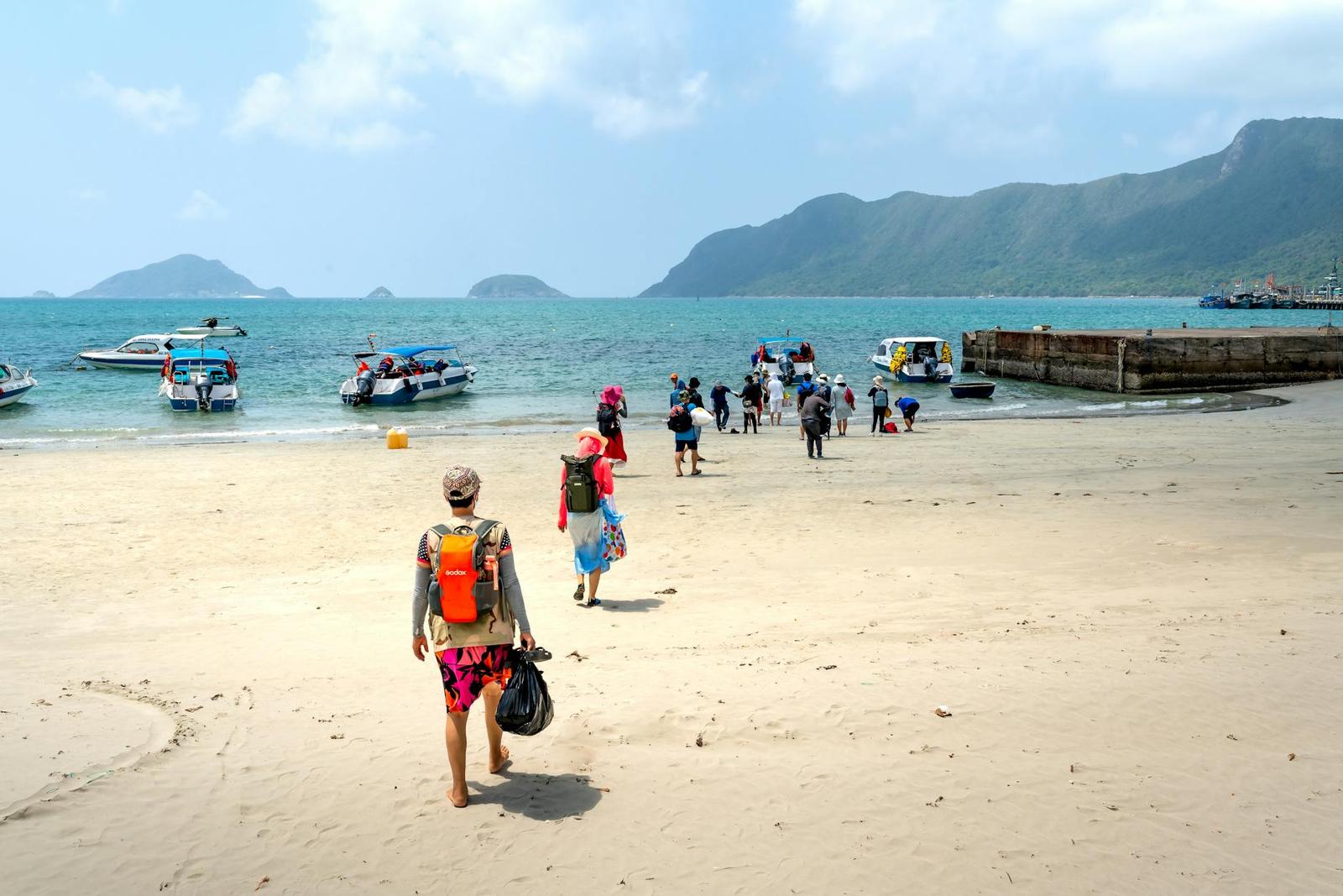 Travellers with backpacks walking along a beach toward moored boats