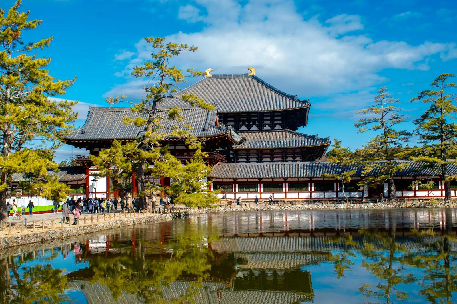 Large wooden temple hall reflected in a still pond with visitors along the shore