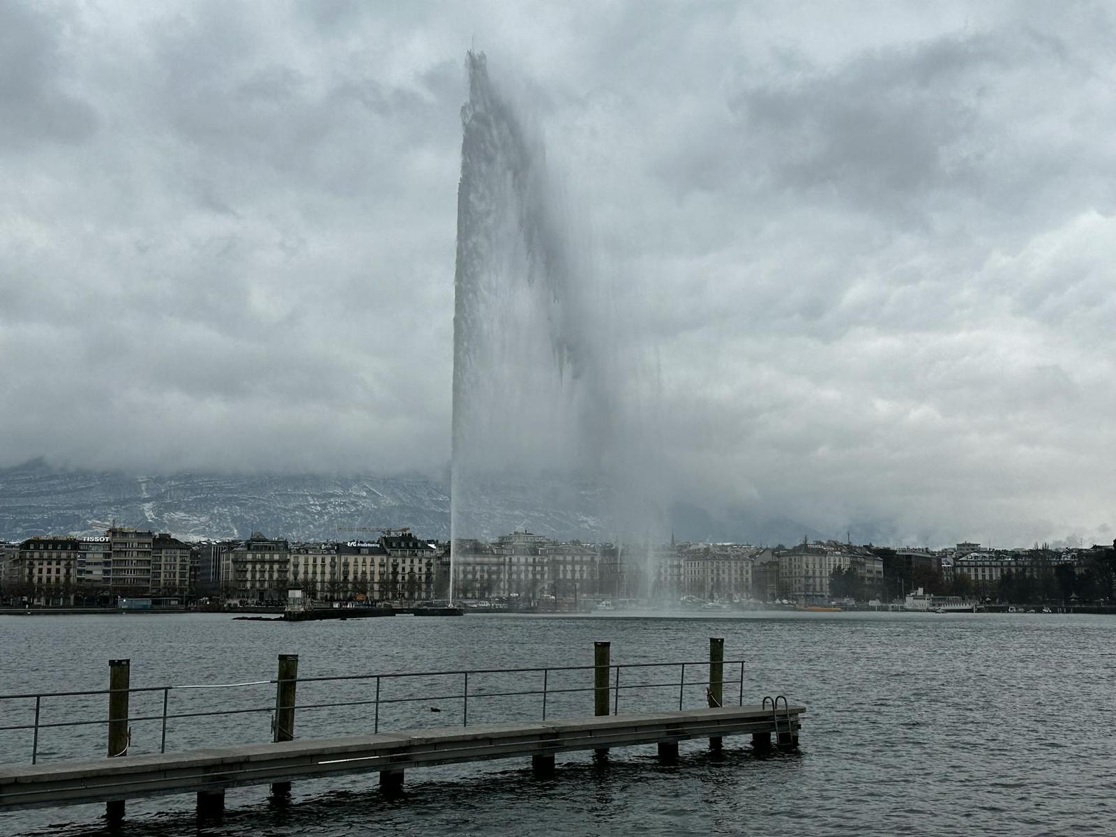 Tall fountain jet spraying over a lake beside a cloudy city skyline