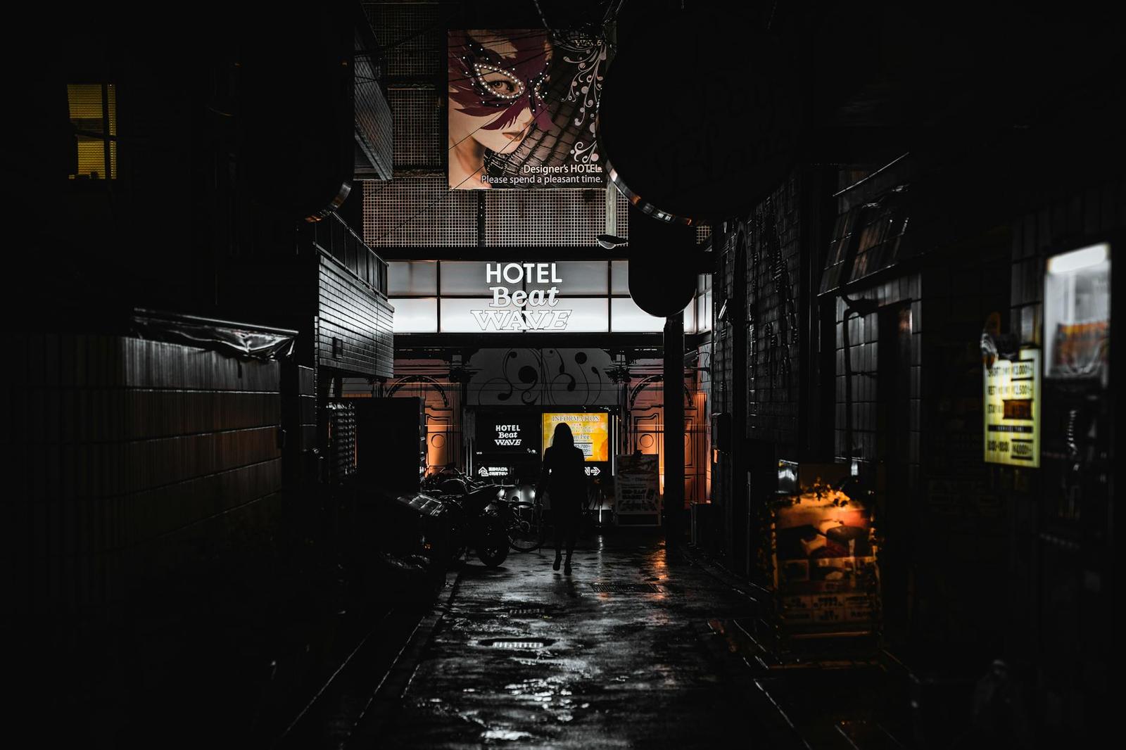 A rain-slicked alley lit by a hotel sign with a figure walking through