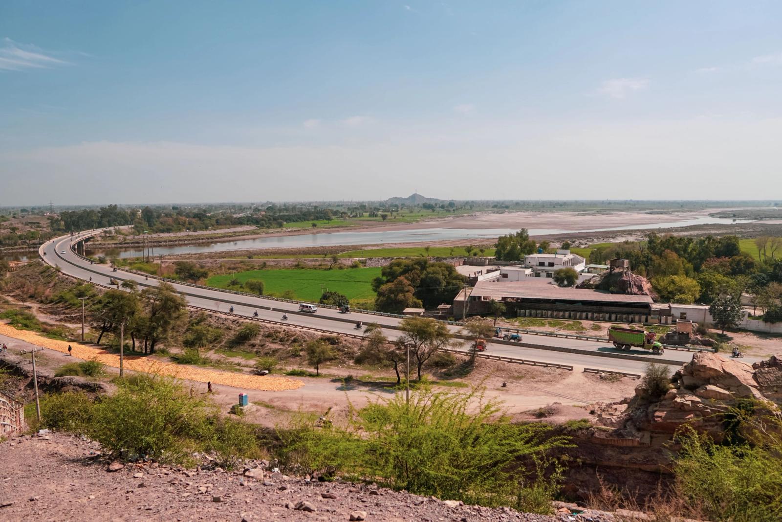 Wide-angle view of highway curving through scenic landscape with river and lush fields under a clear blue sky