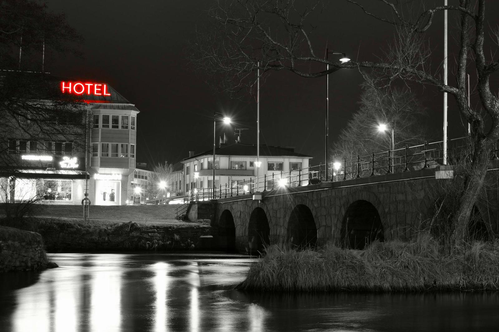 A black and white night scene of a lit HOTEL sign beside a stone bridge over a river