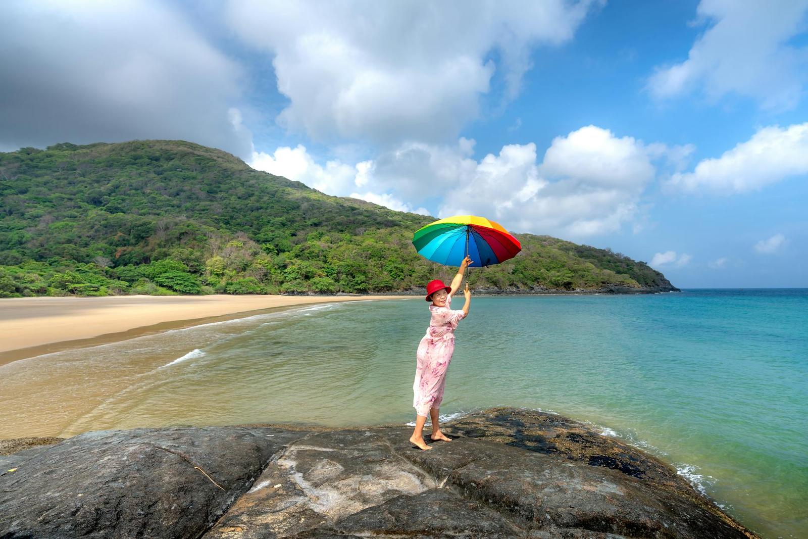 Woman holding a rainbow umbrella on a rock at a forested beach