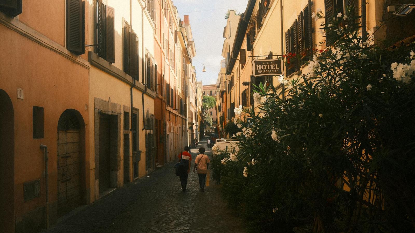 Two people walking down a sunlit cobbled street past a hotel sign and flowering shrubs