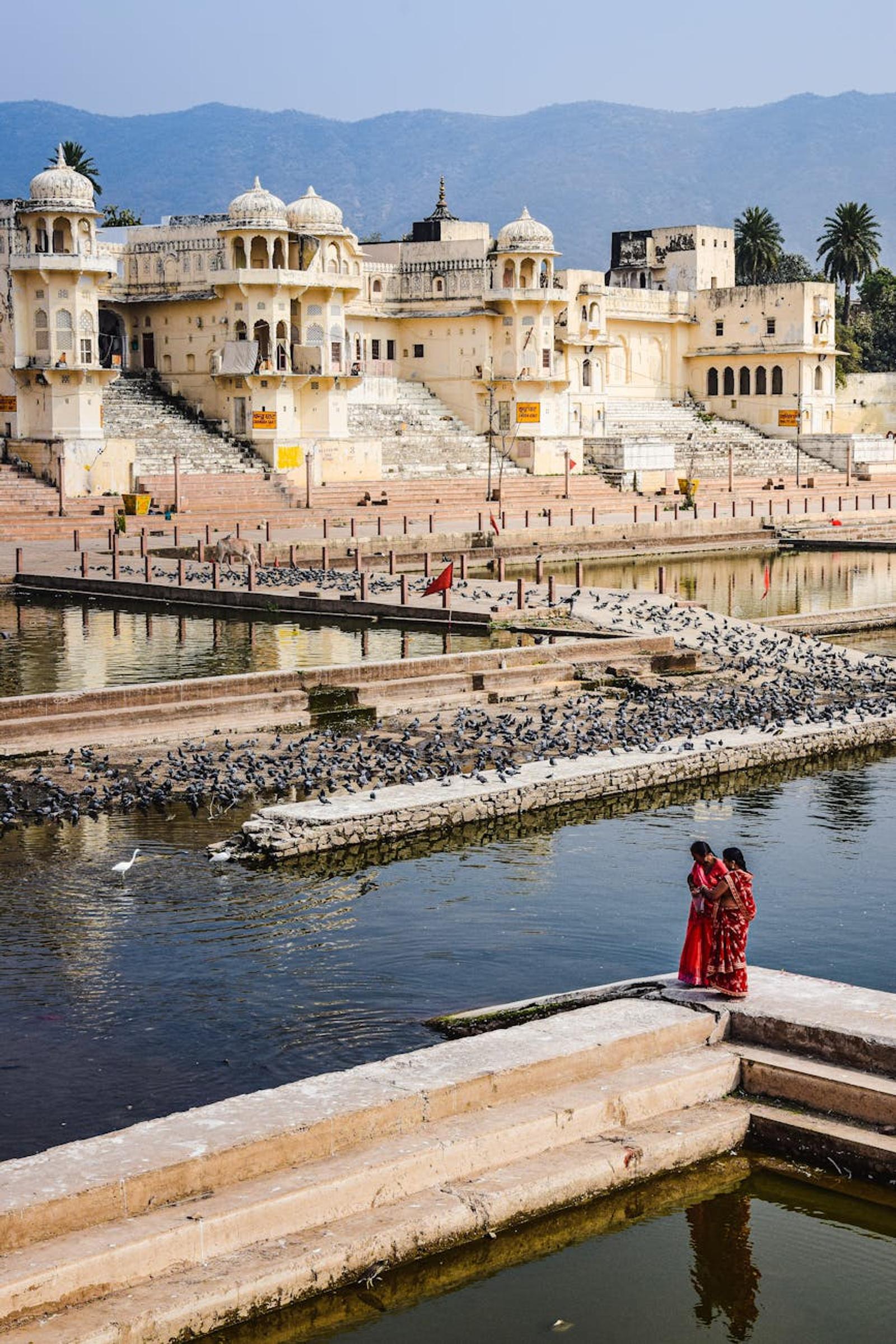 Women in red saris beside stepped lakeside ghats with pigeons and distant hills