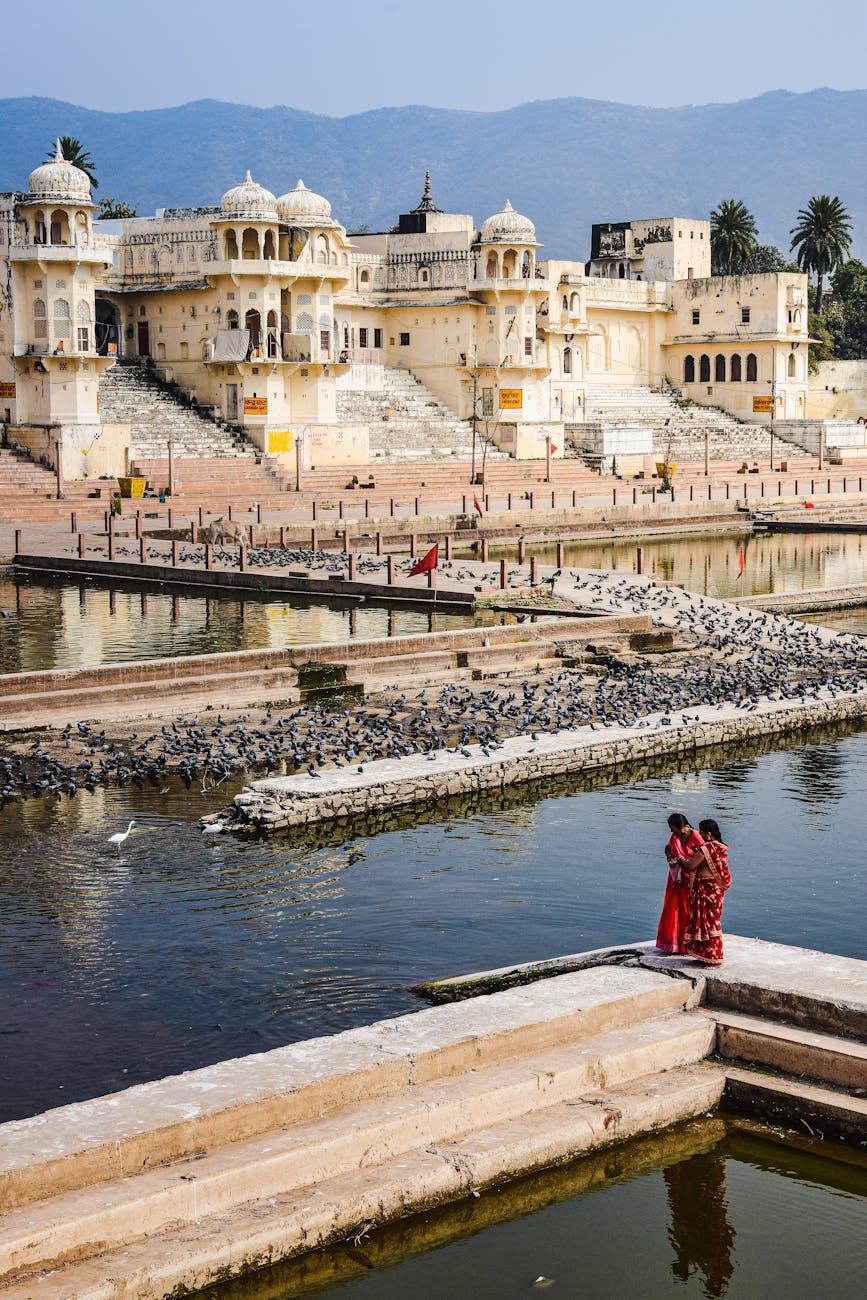 Women in red saris beside stepped lakeside ghats with pigeons and distant hills