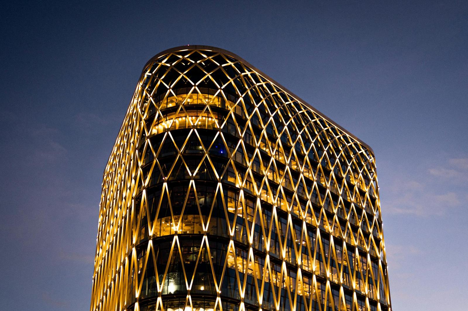 A curved skyscraper with a glowing diamond-lattice facade at dusk